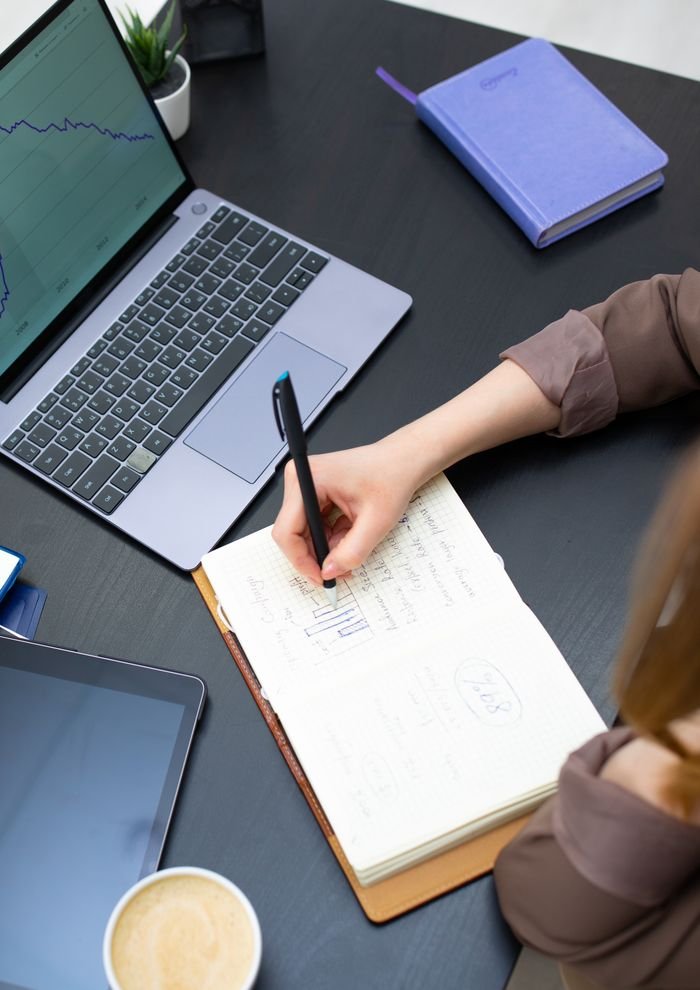 An overhead view of a person writing in a notebook at a desk with a laptop and coffee.